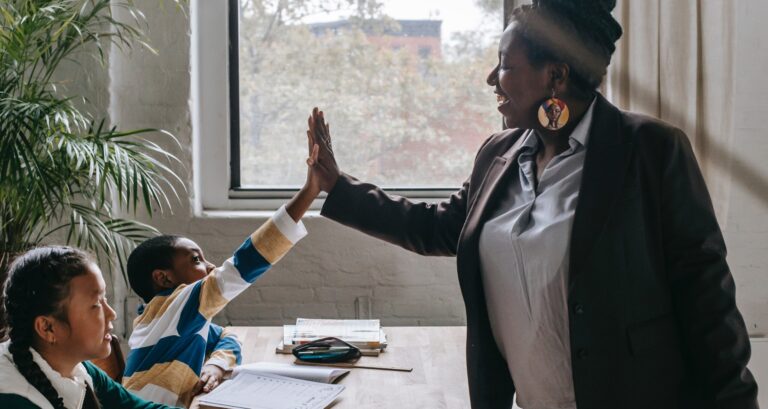 Older African American teacher gives younger African American young boy sitting at a desk a high five. A young Asian girl looks towards them. The diversity is emphasized by having an African American teacher while diverse students.